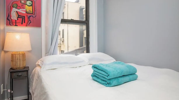 A neatly made bed with white linens and a teal towel, a lamp on a side table, and a colorful abstract painting on the wall. - Modern 4-Bedroom Stay in the Heart of Lenox Hill! (New York)