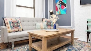 A living room with a grey sofa, colorful cushions, a wooden coffee table, a painting, and a TV mounted on the wall. - Modern 4-Bedroom Stay in the Heart of Lenox Hill! (New York)