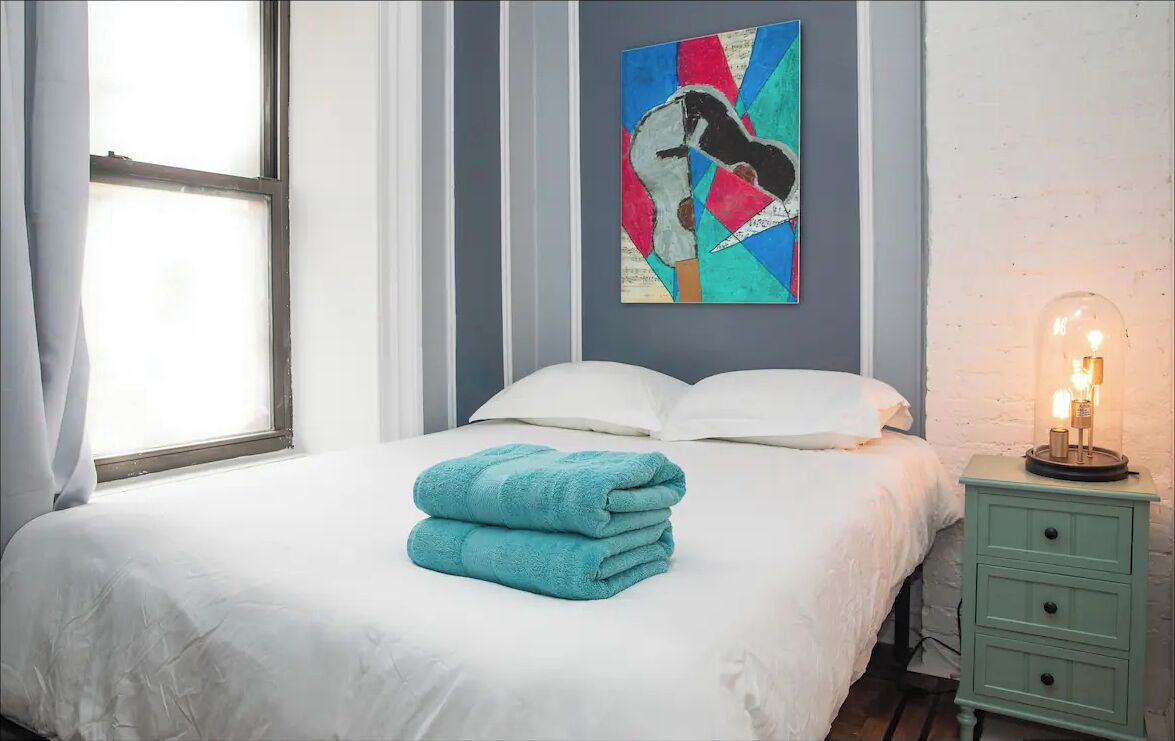 A neatly made bed with white linens and a stack of teal towels, a wall-mounted abstract painting, a bedside table with a lamp, and a window with curtains.