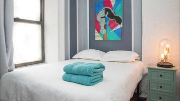 A neatly made bed with white linens and a stack of teal towels, a wall-mounted abstract painting, a bedside table with a lamp, and a window with curtains.