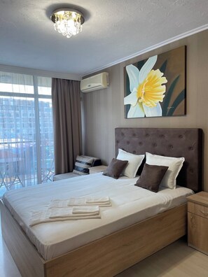 A modern hotel room with a large bed, a ceiling light, an air conditioning unit, a painting of a flower, and a view of the city through large windows. - 2-bedroom apartment in amazing Sunny Beach,beachfront location,sea views (Sunny Beach)