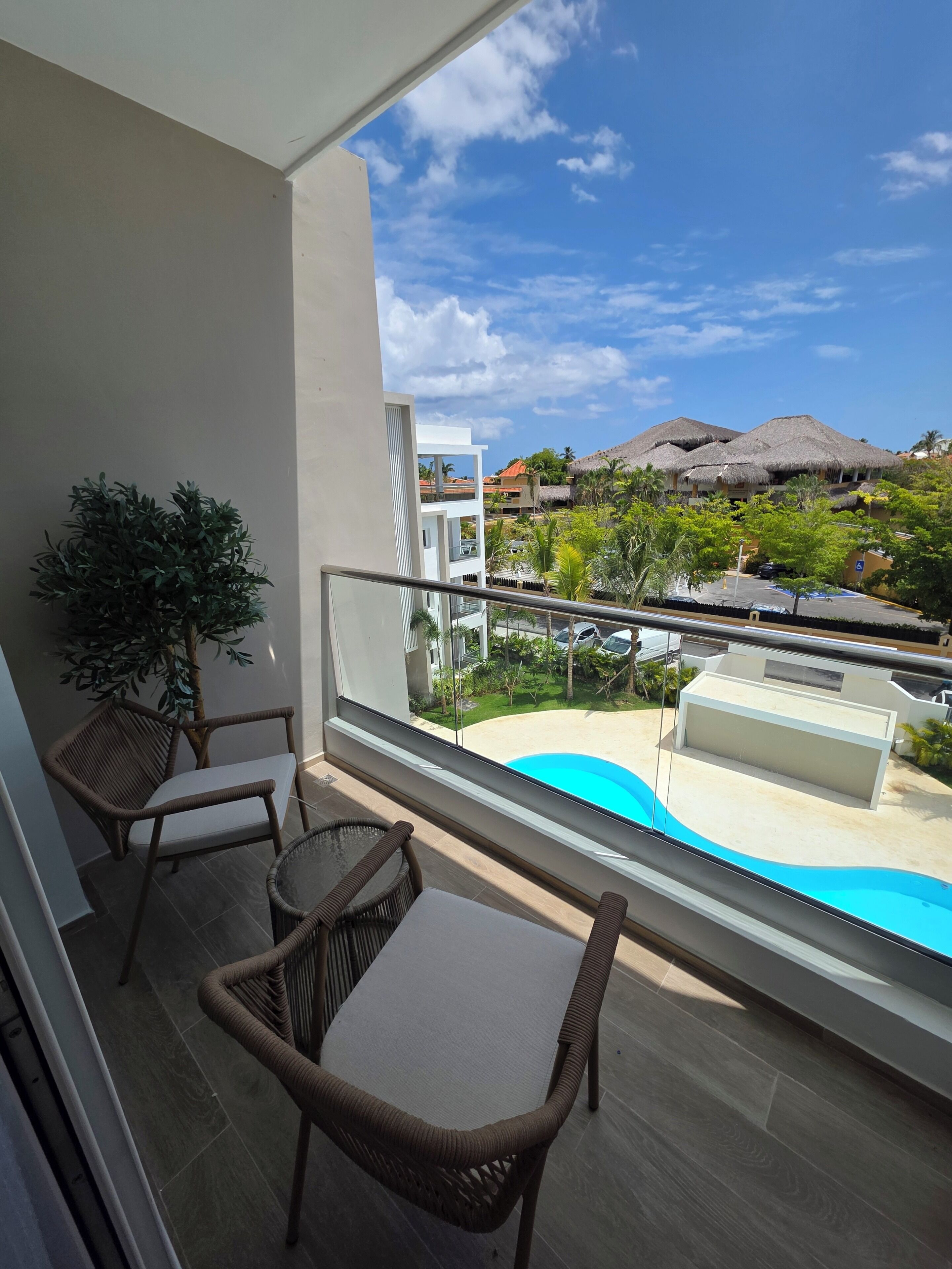 A balcony with wicker furniture, a view of a swimming pool, and buildings with thatched roofs in the background.