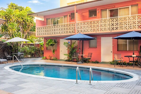 Ein Schwimmbad mit Holzterrasse, ein rotes Gebäude mit Balkon und Gartenmöbeln.