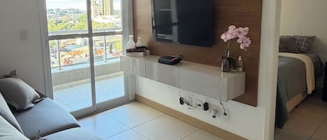 A modern living room with a flat-screen TV mounted on a wooden wall, a sofa, and a bed with a view of the city.