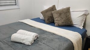 Een net opgemaakt bed met een grijze dekbedovertrek, twee bruine kussens en een wit kussen. Een opgevouwen witte handdoek ligt op het bed.