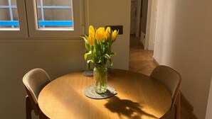 A round wooden dining table with four chairs, a vase of yellow flowers, and a window with blinds. - Stunning Designer Apartment in the Heart of Paris (11th Arrondissement) (Paris)