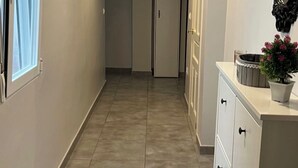 Un couloir avec un sol carrelé, une porte au fond et une coiffeuse agrémentée d’un miroir et d’une plante en pot.