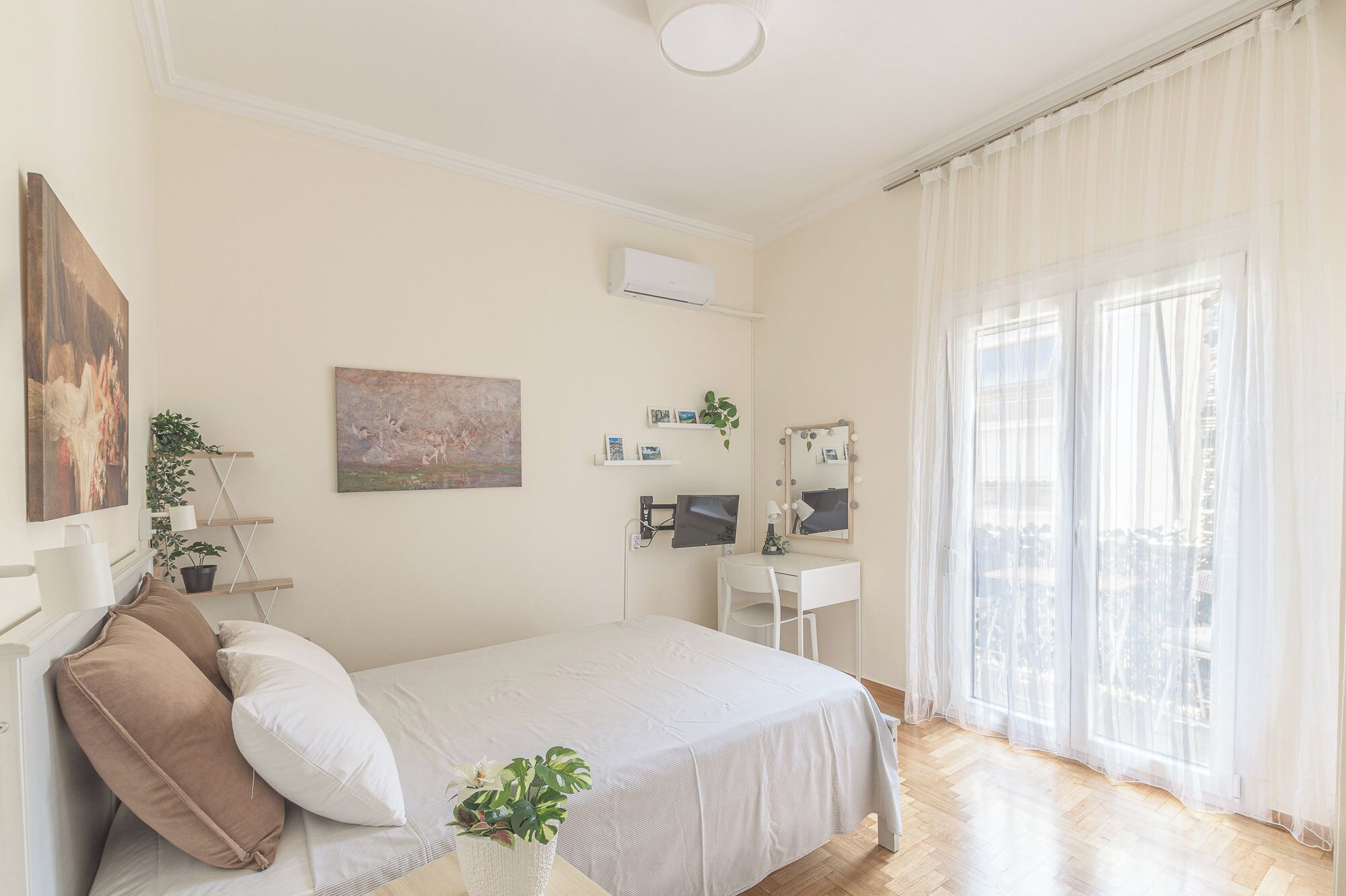 A bedroom with a bed, a desk with a mirror, a window with sheer curtains, and wall-mounted shelves with plants.