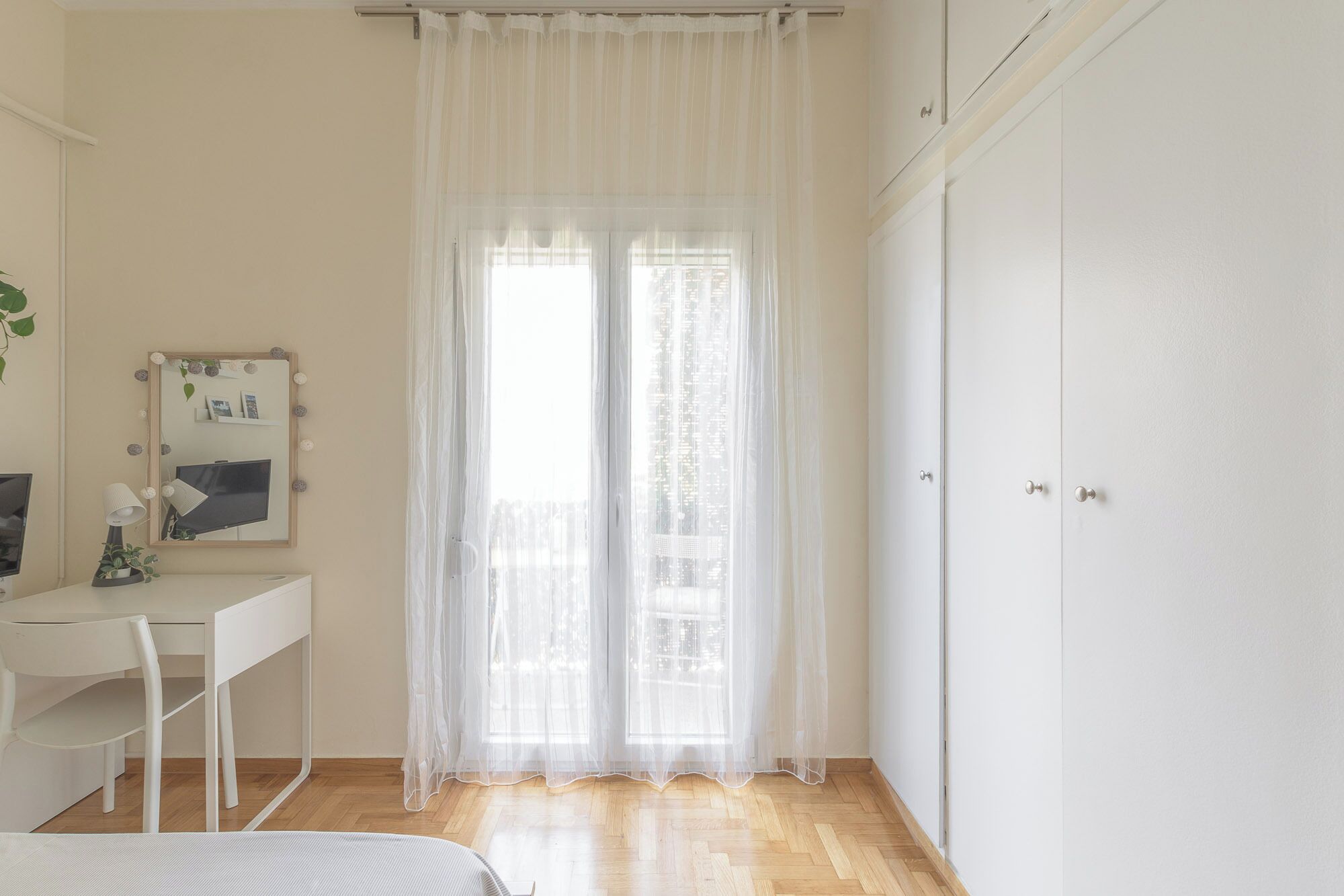 Une pièce comprenant un bureau blanc, une chaise, un miroir avec éclairage intégré, une fenêtre avec des rideaux légers et une armoire.