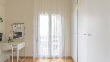 Une pièce comprenant un bureau blanc, une chaise, un miroir avec éclairage intégré, une fenêtre avec des rideaux légers et une armoire.