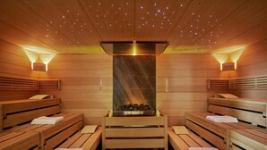 Un sauna avec des bancs en bois, un foyer central et des luminaires fixés aux murs.