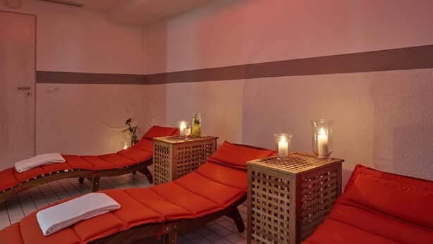 A spa room with three massage tables, each with a red cushion and a towel. There are two wooden side tables with candles and bottles. - Sunday Hotel Köln Hürth Disability Access (Hürth)