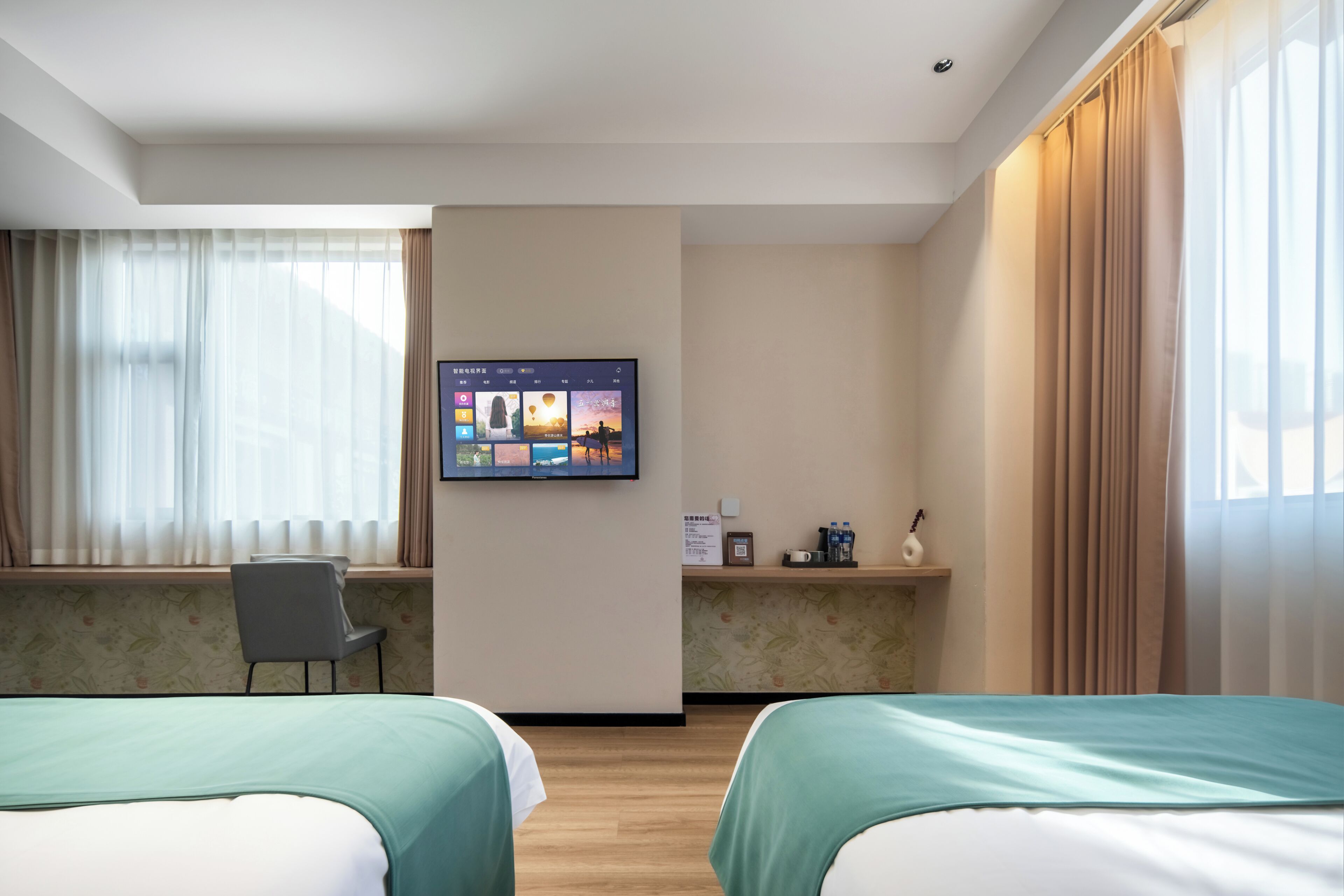 A hotel room with two beds, a desk, a TV on the wall, and a window with curtains.