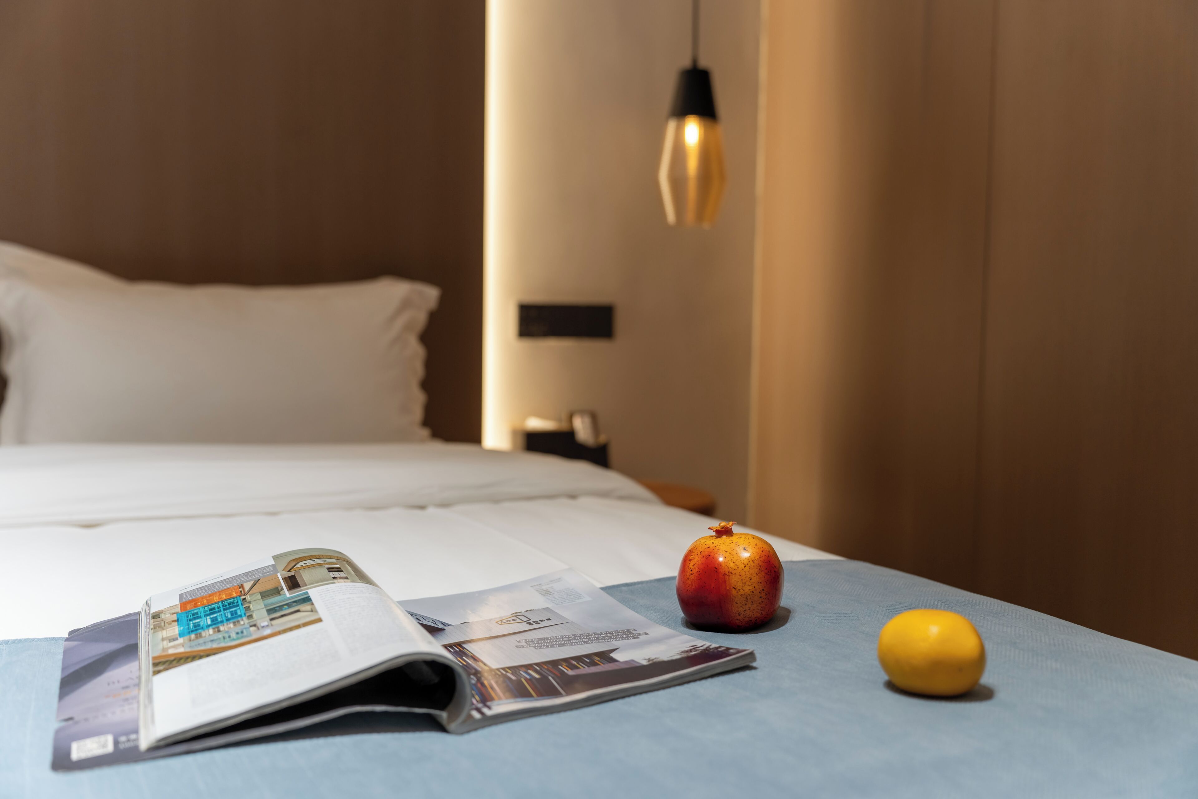Una cama bien tendida con una colcha blanca y almohadas, una revista sobre la cama, una granada y un limón sobre un mantel azul.
