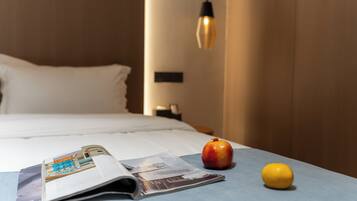 Una cama bien tendida con una colcha blanca y almohadas, una revista sobre la cama, una granada y un limón sobre un mantel azul.