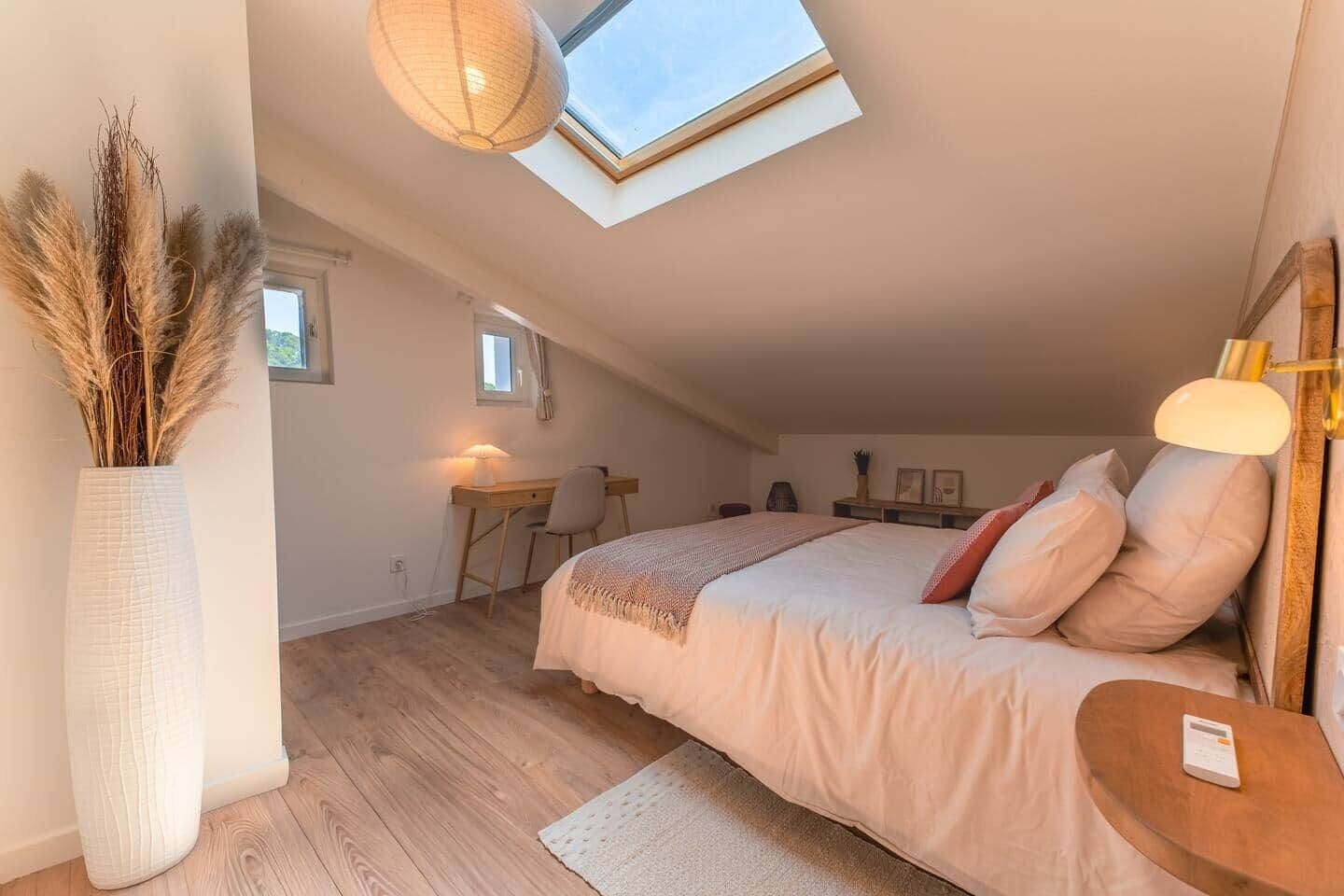 A bedroom with a skylight, a desk, a chair, a bed with pillows, and a bedside table with a lamp.