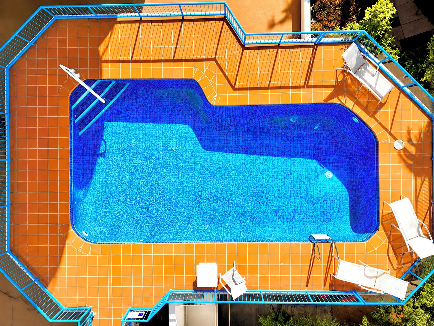 Une piscine rectangulaire avec une terrasse carrelée, entourée d’une rambarde en métal bleu, et équipée de chaises longues blanches et d’un transat.
