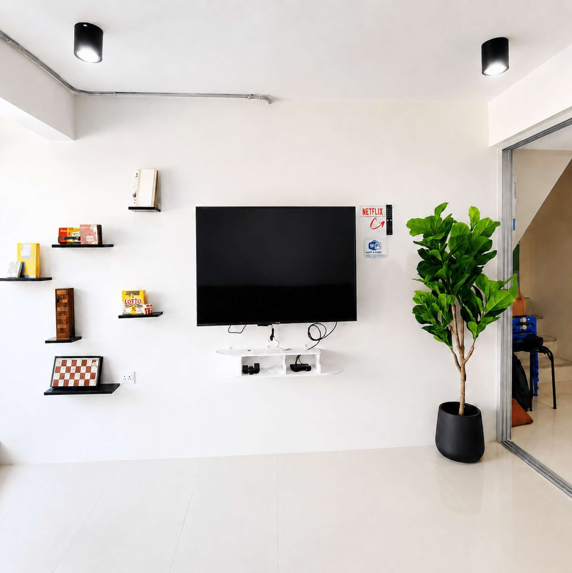 A modern living room with a flat-screen TV mounted on the wall, surrounded by shelves with various items, a potted plant, and a visible Wi-Fi sign.