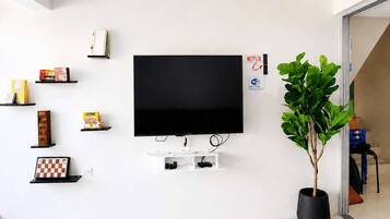 A modern living room with a flat-screen TV mounted on the wall, surrounded by shelves with various items, a potted plant, and a visible Wi-Fi sign.