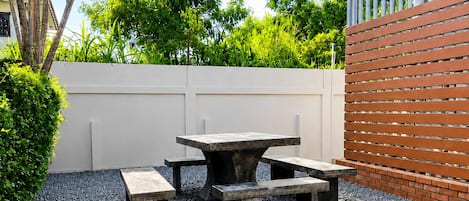 Un patio al aire libre con una mesa y bancos, rodeado por un muro blanco y una valla de ladrillo.