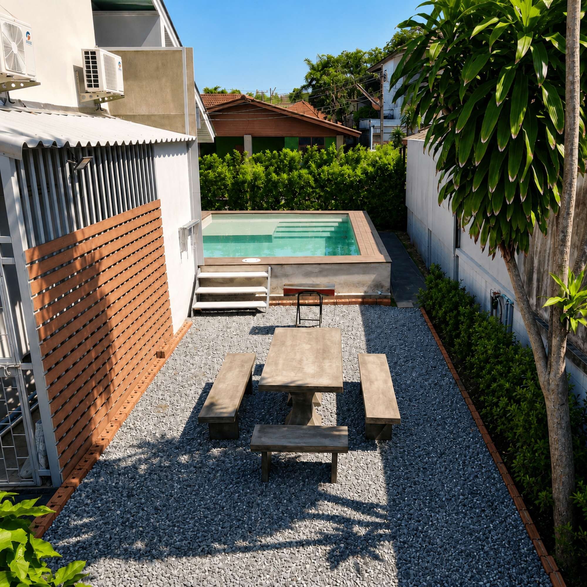 A rectangular swimming pool with a wooden deck and benches, surrounded by a gravel area and a low brick wall.