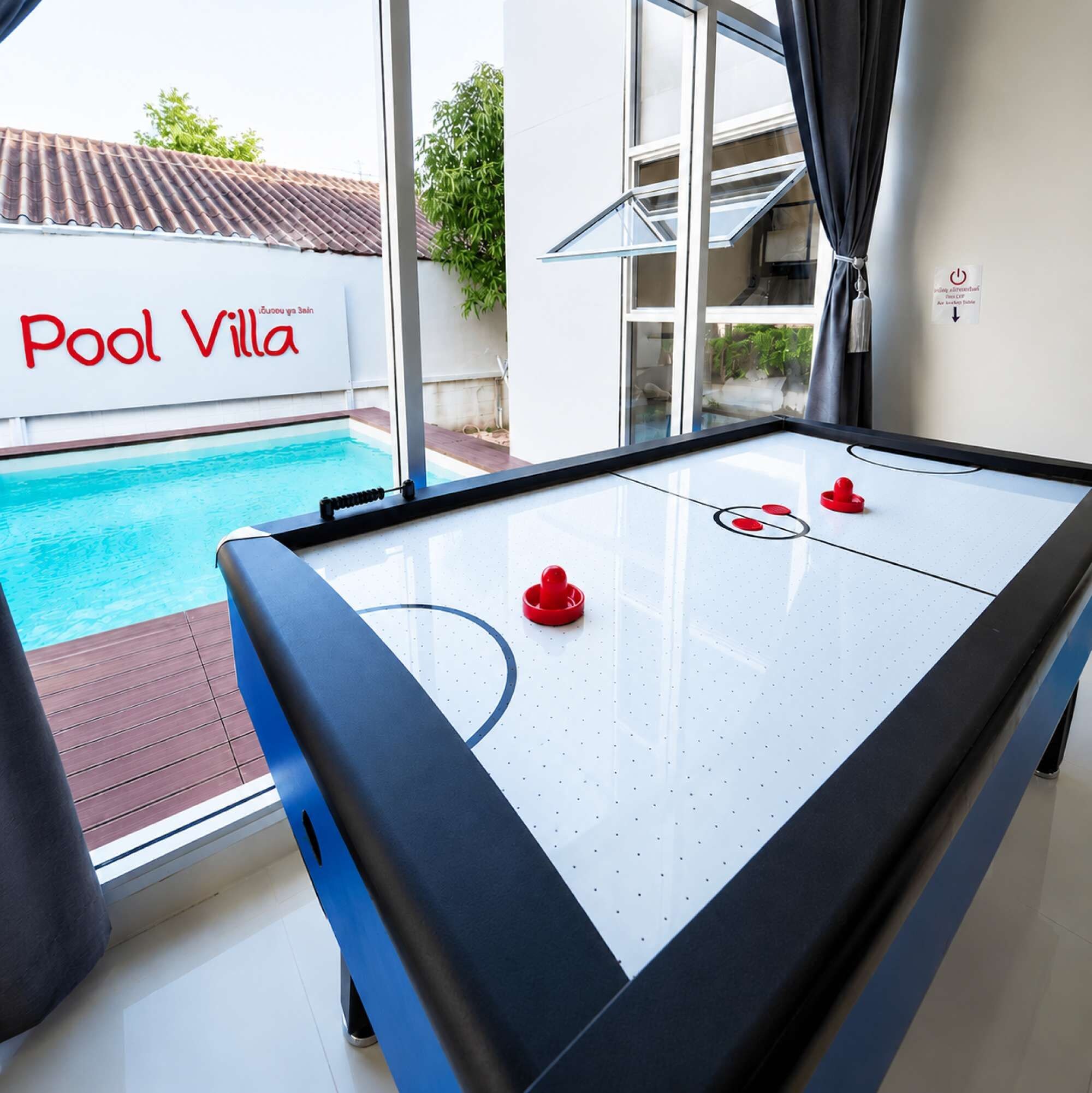 A pool table with a clear view of a swimming pool outside, a sign reading 'Pool Villa', and a window with curtains.
