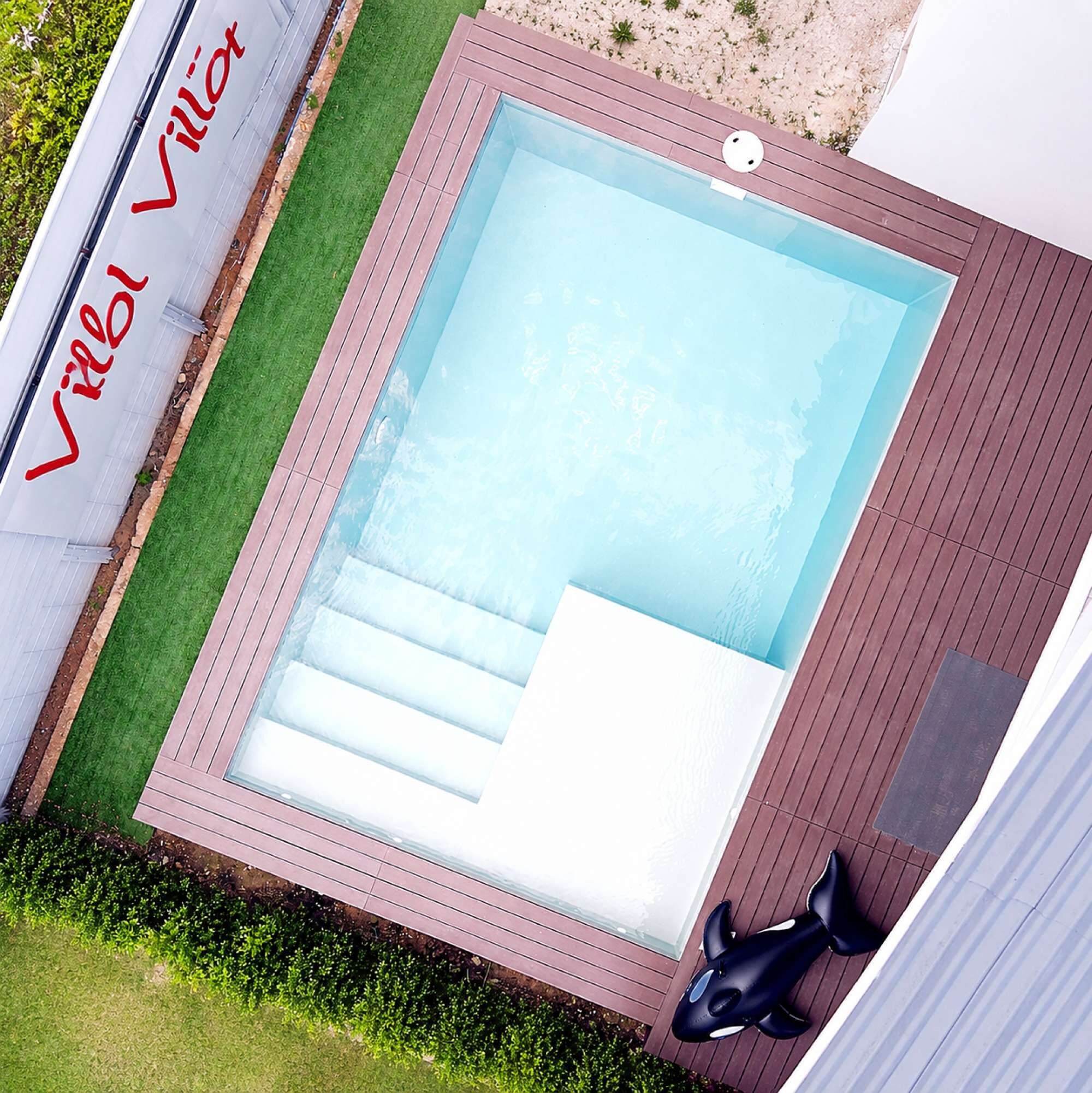 A rectangular swimming pool with a wooden deck surrounding it, a small inflatable orca, and a sign with red lettering.