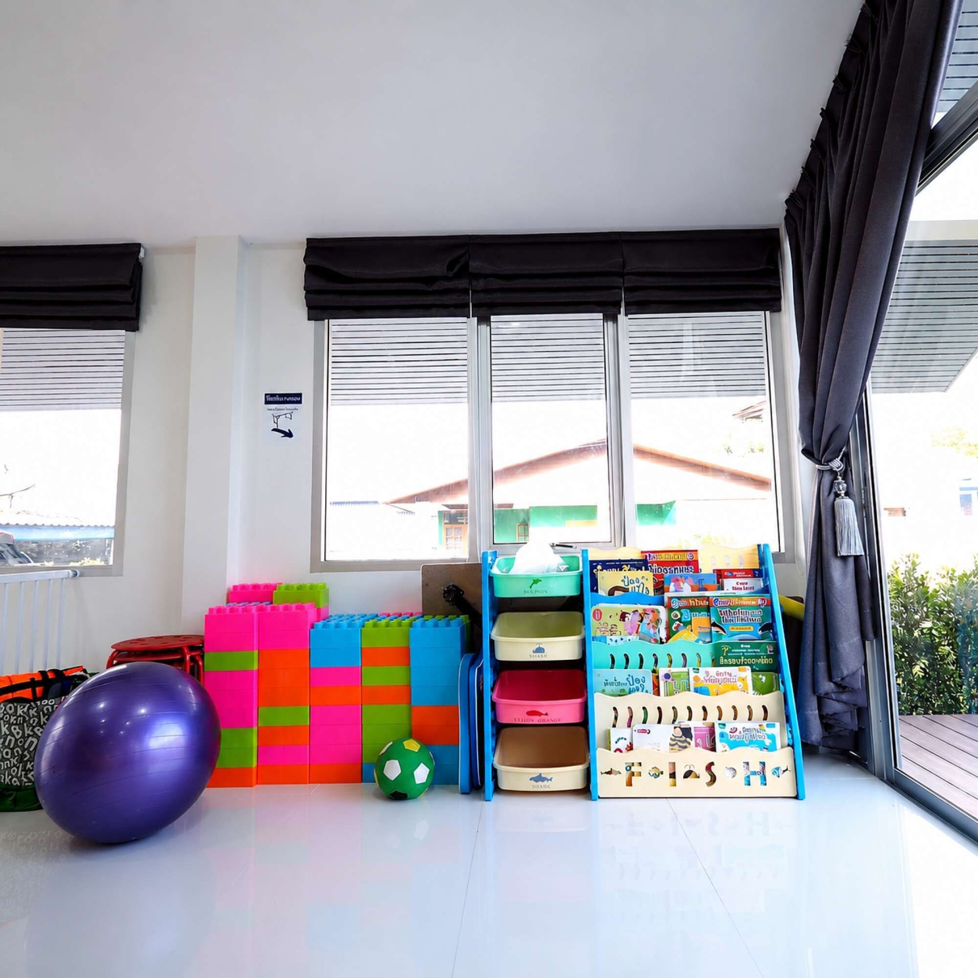 Una habitación con ventanas amplias, un área de juegos con bloques de construcción de colores, una pelota de ejercicios morada y un estante con libros infantiles.