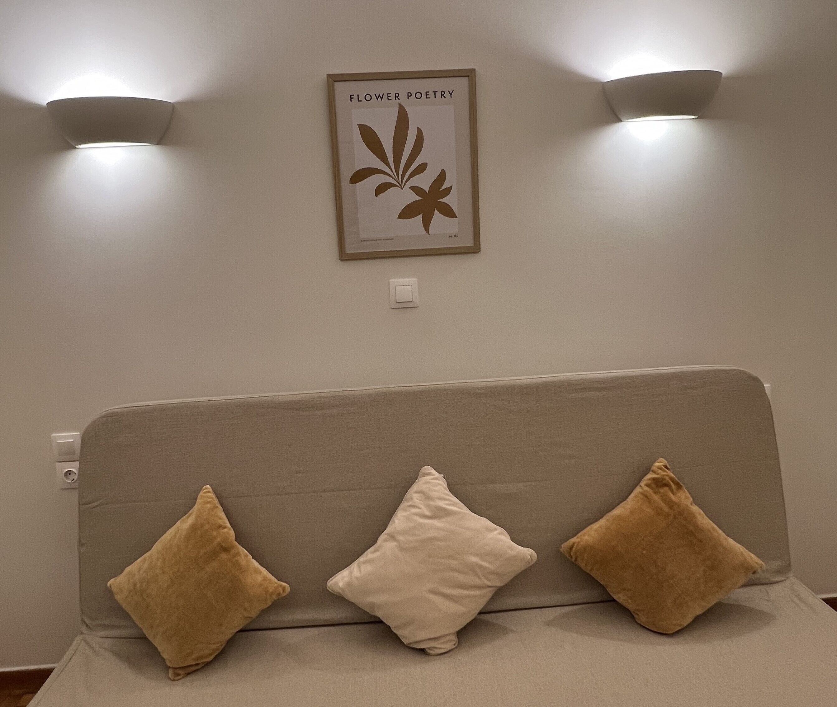 Ein sofasitz in neutralen Farbtönen mit drei Kissen, darüber ein gerahmtes Kunstwerk mit dem Titel 'Flower Poetry' und je eine Wandleuchte links und rechts davon.