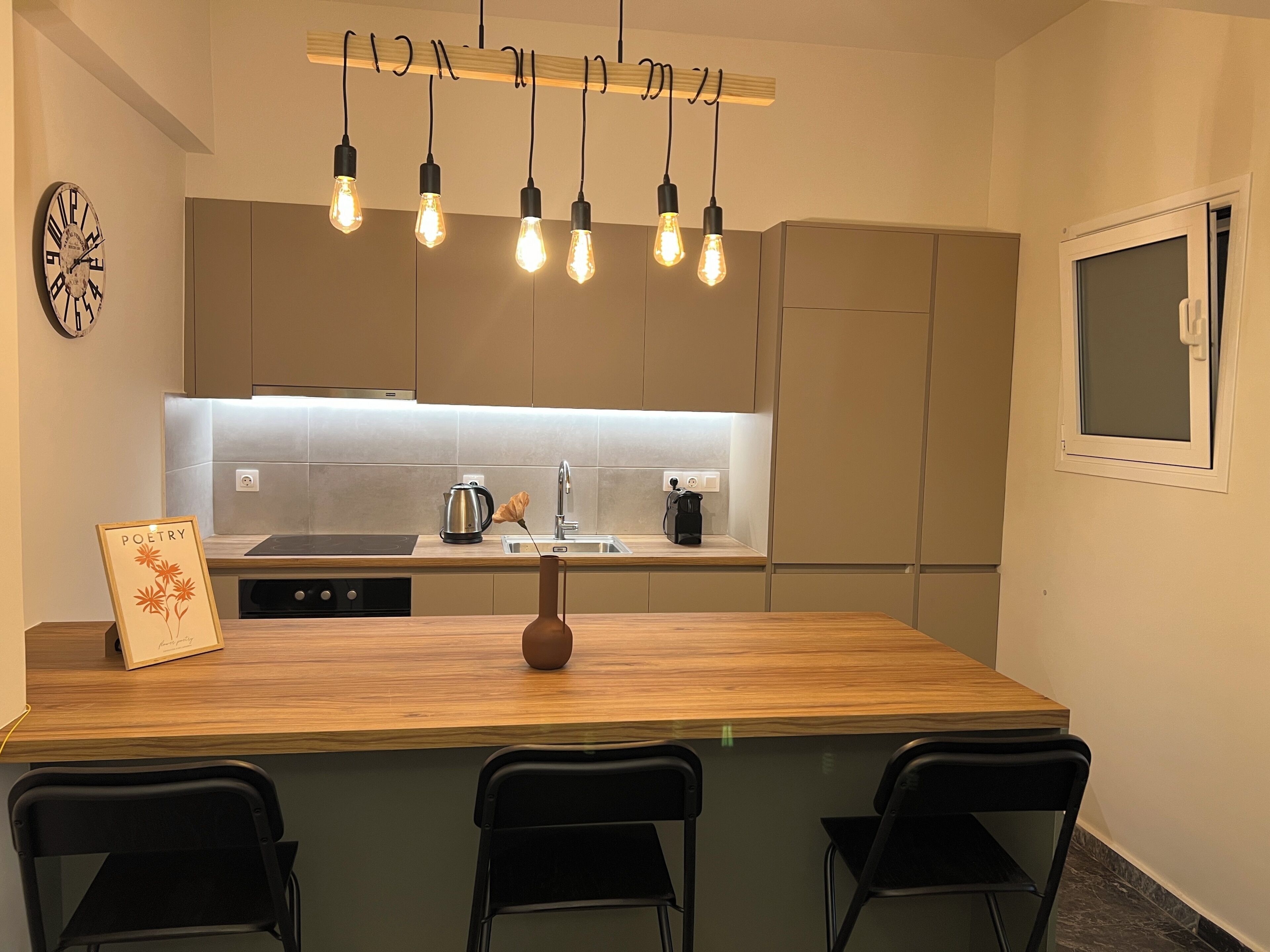 A modern kitchen with a wooden island, black chairs, a clock, and a sign reading 'POETRY'.