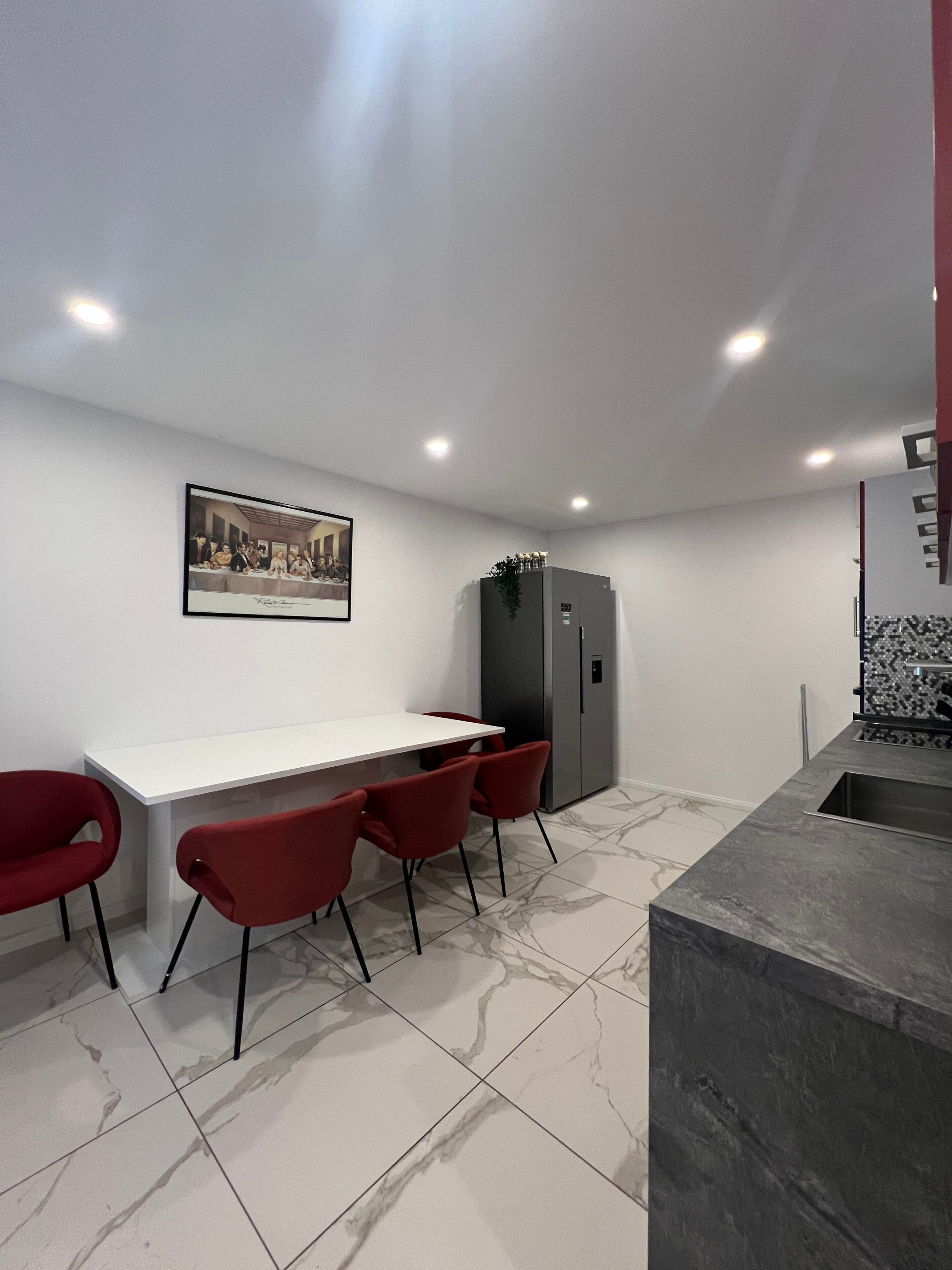 Una cucina moderna con un piano di lavoro in marmo, un frigorifero e un'area pranzo con un tavolo bianco e sedie rosse.