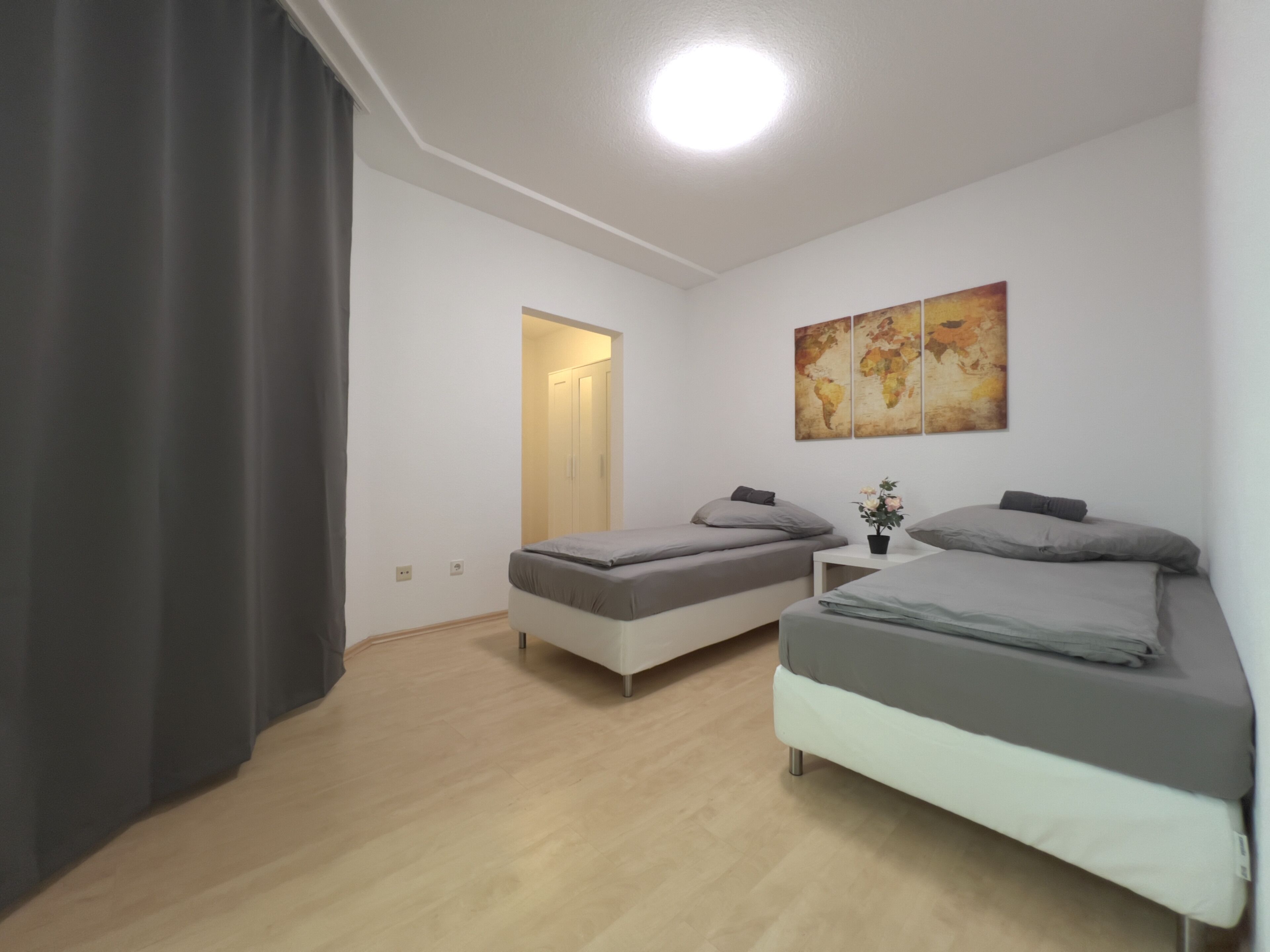 Una habitación con dos camas, una mesita con una planta, un mapa del mundo en la pared y una puerta que da a otra habitación.