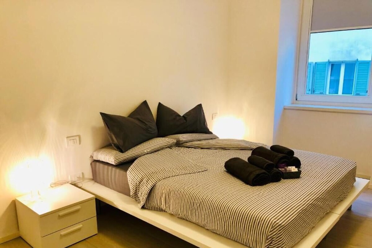 Ein Bett mit gestreifter Bettdecke, zwei Kissen und zusammengerollten Handtüchern auf einem weißen Nachttisch.