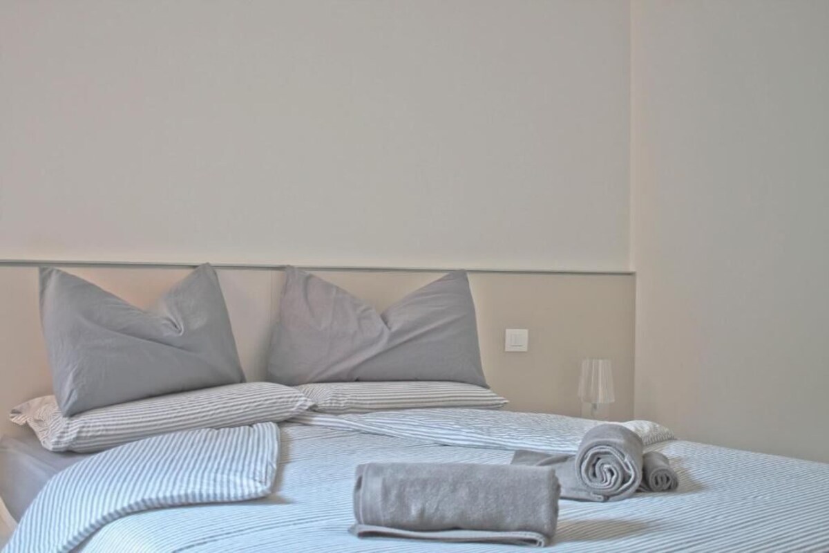 Ein ordentlich bezogenes Bett mit Kissen und einem zusammengerollten Handtuch, eine Nachttischlampe und eine schlichte Wand.
