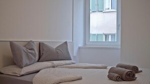 Ein Bett mit gestreifter Bettwäsche und Kissen, ein Fenster mit Blick auf Gebäude und zusammengerollte Handtücher auf dem Bett.
