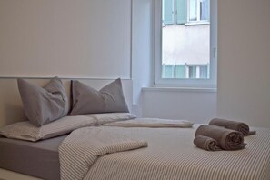 Ein Bett mit gestreifter Bettwäsche und Kissen, ein Fenster mit Blick auf Gebäude und zusammengerollte Handtücher auf dem Bett.