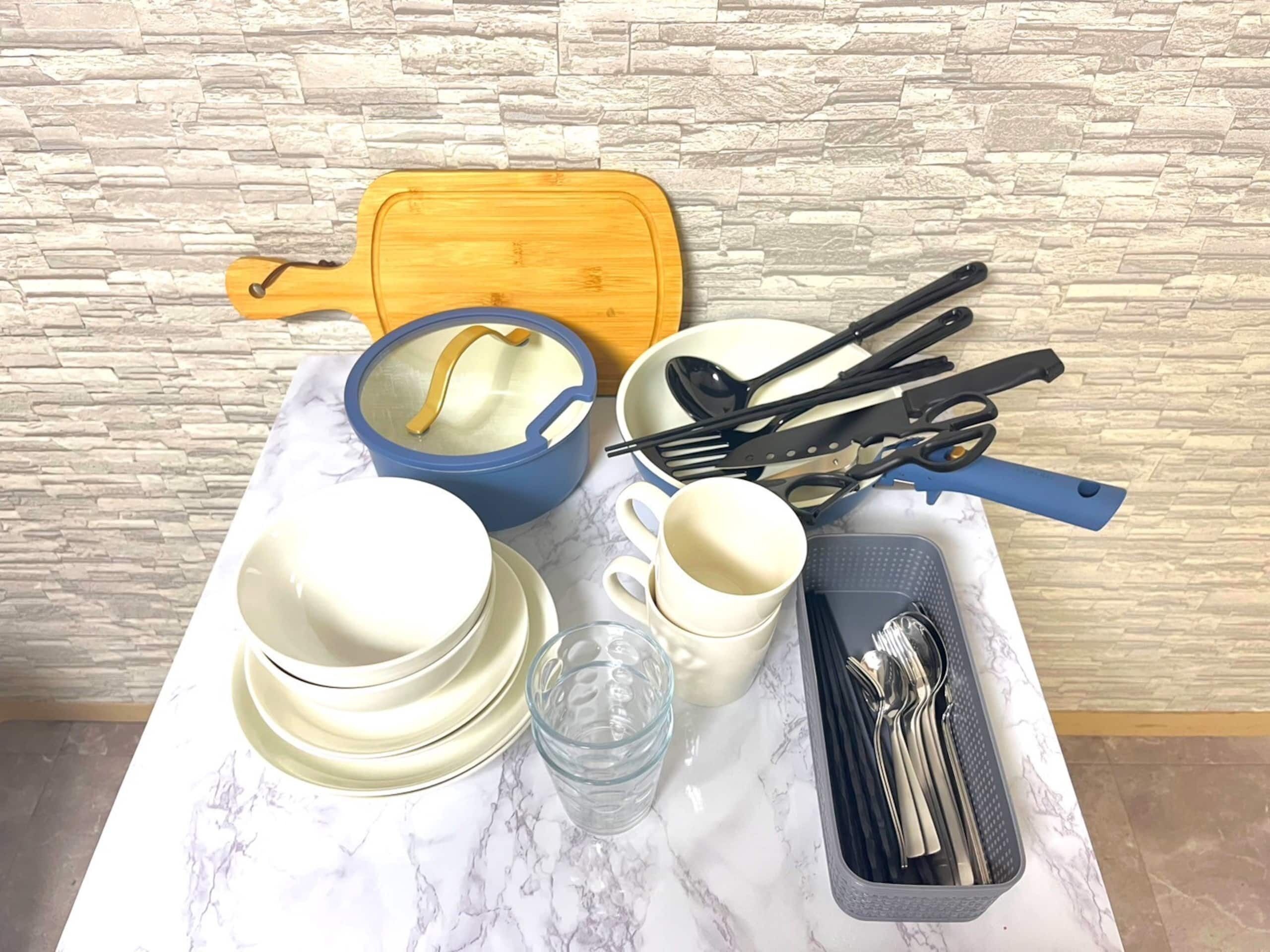 Uma mesa de mármore com um vaso azul, uma tábua de corte de madeira, um copo, uma xícara branca, um conjunto de utensílios pretos e um tabuleiro cinzento.