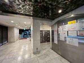 A hallway with a tiled floor, a metal wall with informational signs, and a staircase in the background. - Dainess Ichibankan Shibuya (Shibuya-ku)