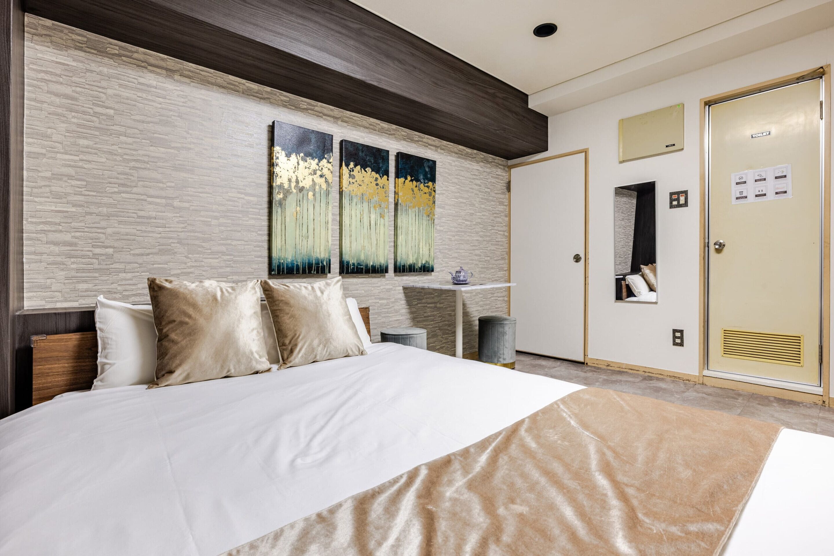 A hotel room with a large bed, a painting on the wall, a small table with a vase, and a wardrobe with a toilet sign.