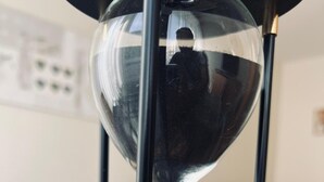 Un reloj de arena con estructura de vidrio transparente, sostenido por soportes negros, sobre una superficie con un cable y papeles al fondo.