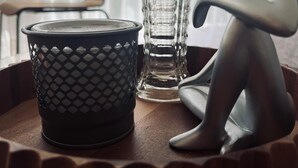 Una mesa con una cesta negra texturizada, un florero de vidrio transparente y una escultura minimalista.