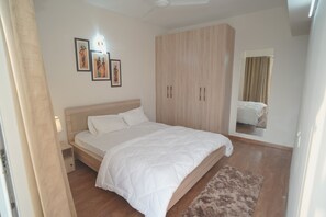 A bedroom with a bed, wooden wardrobe, bedside table, and wall-mounted artwork. - 2-bedroom apartment in peaceful Greater Noida with AC and gym access (GREATER NOIDA)