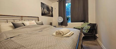 Un dormitorio con cama, ventilador de pared, ventana con cortinas, radiador y un cuadro.