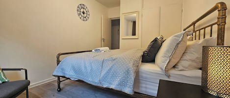 A bedroom with a bed, a chair, a bedside table, a clock, and a mirror on the wall.