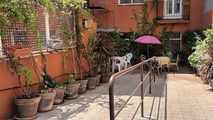 Un corridoio esterno stretto con piante in vaso su entrambi i lati, una ringhiera in metallo e un tavolo con sedie sotto un ombrello rosa.
