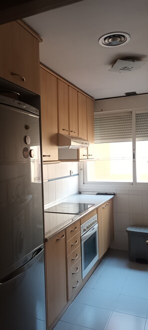 A kitchen with wooden cabinets, a stainless steel refrigerator, a built-in oven, and a window with blinds. - Apartment for medium- to long-term stays in Malasaña (Madrid)