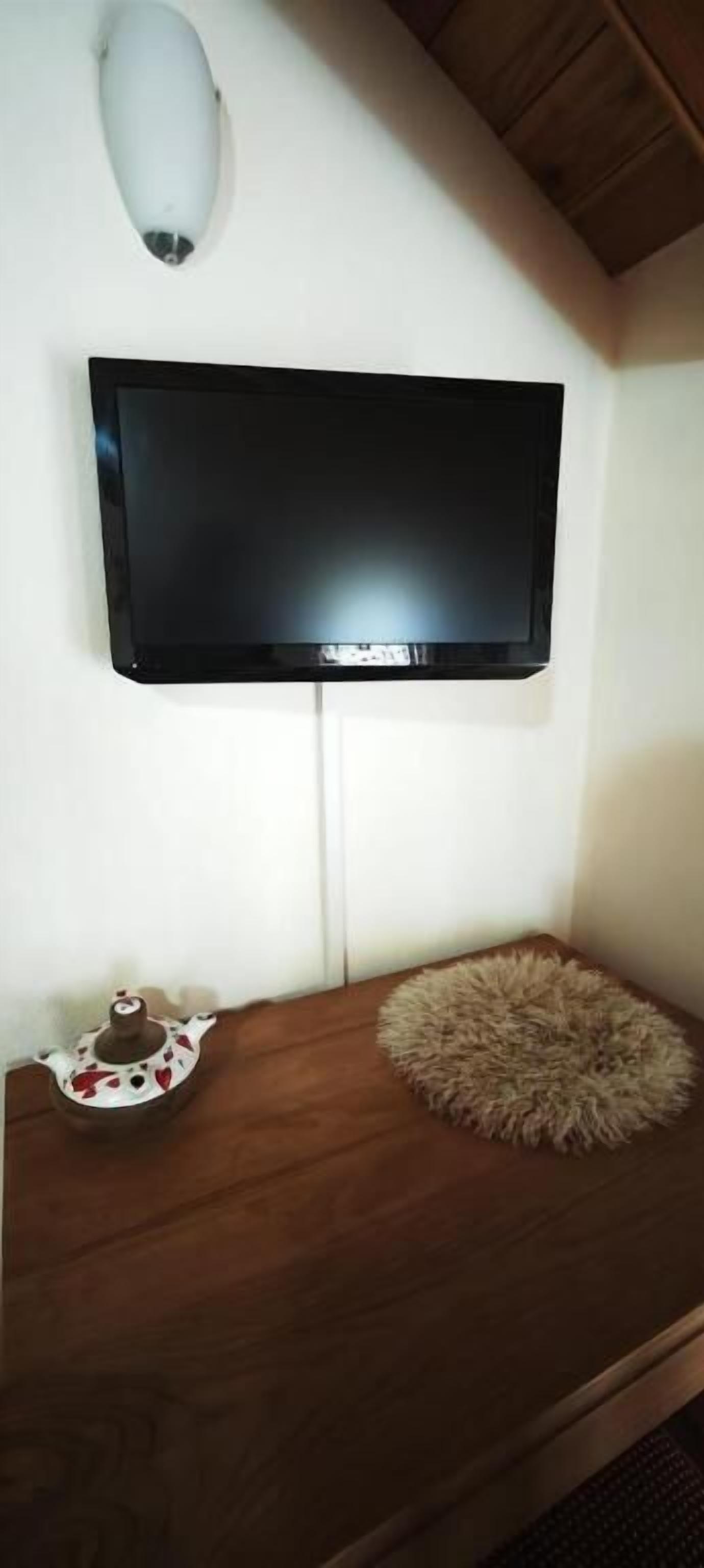 Un televisor de pantalla plana montado en la pared, un mueble de madera con un rincón decorativo que incluye una tetera y un cojín mullido, y una pared blanca.