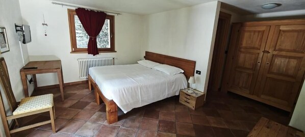 A bedroom with a bed, a wooden nightstand, a chair, a radiator, a window with curtains, and a wardrobe. - Affittacamere I PICCHI (Gressan)