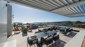 An outdoor patio with a view of the sea, featuring a grill, seating, and a pergola.