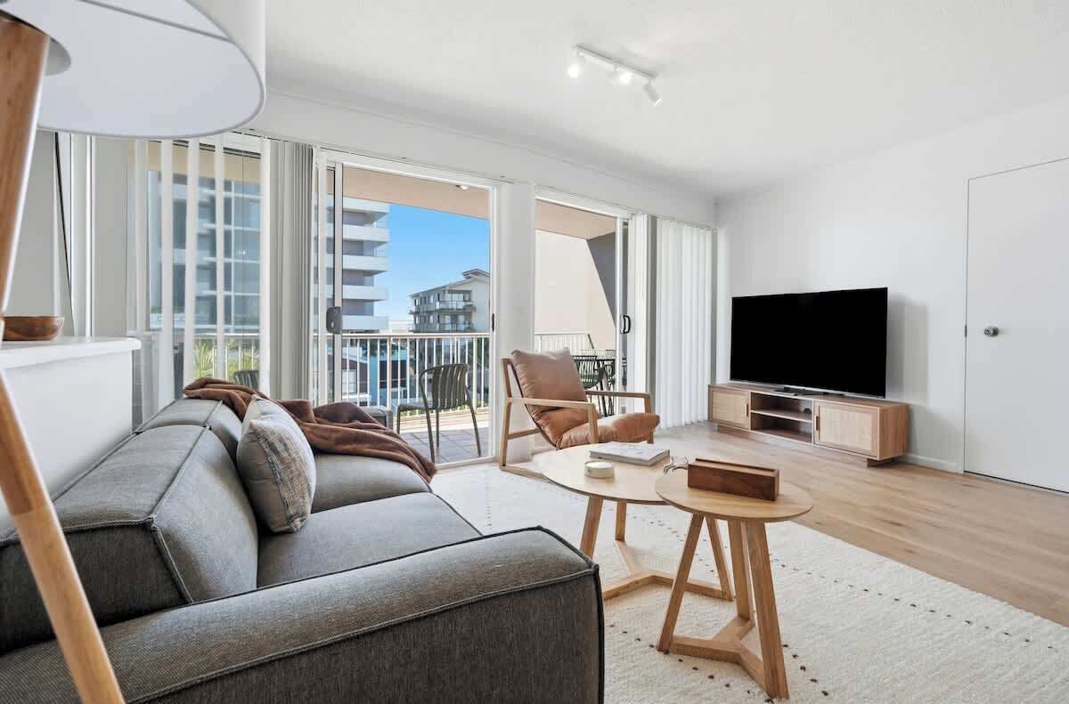 A modern living room with a sofa, a coffee table, a TV, and a balcony with a view of buildings.